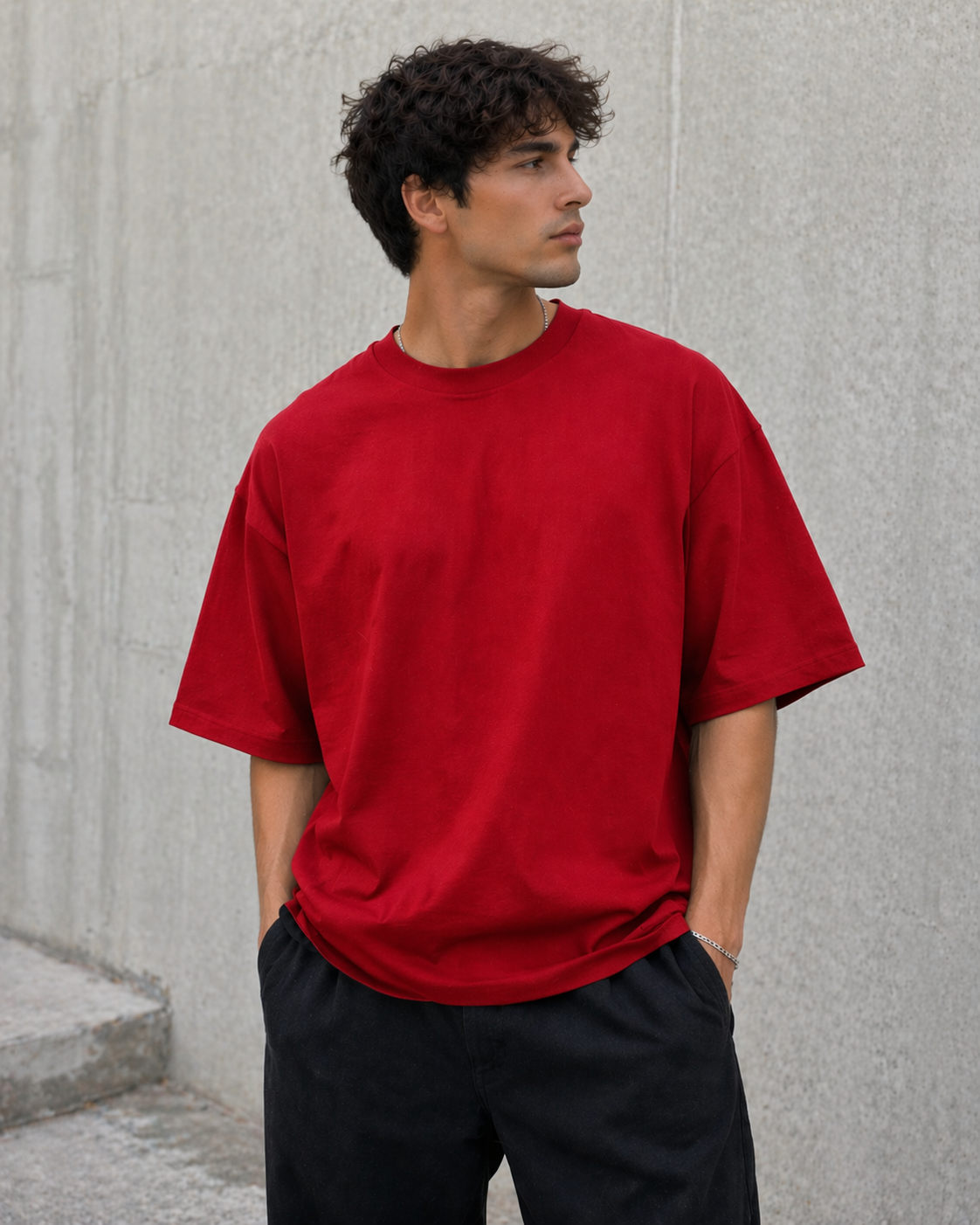 Man wearing a red t-shirt and black pants standing against a gray concrete wall.