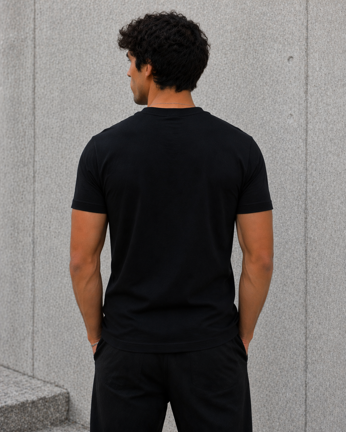 Man wearing a black t-shirt and pants standing against a gray wall.